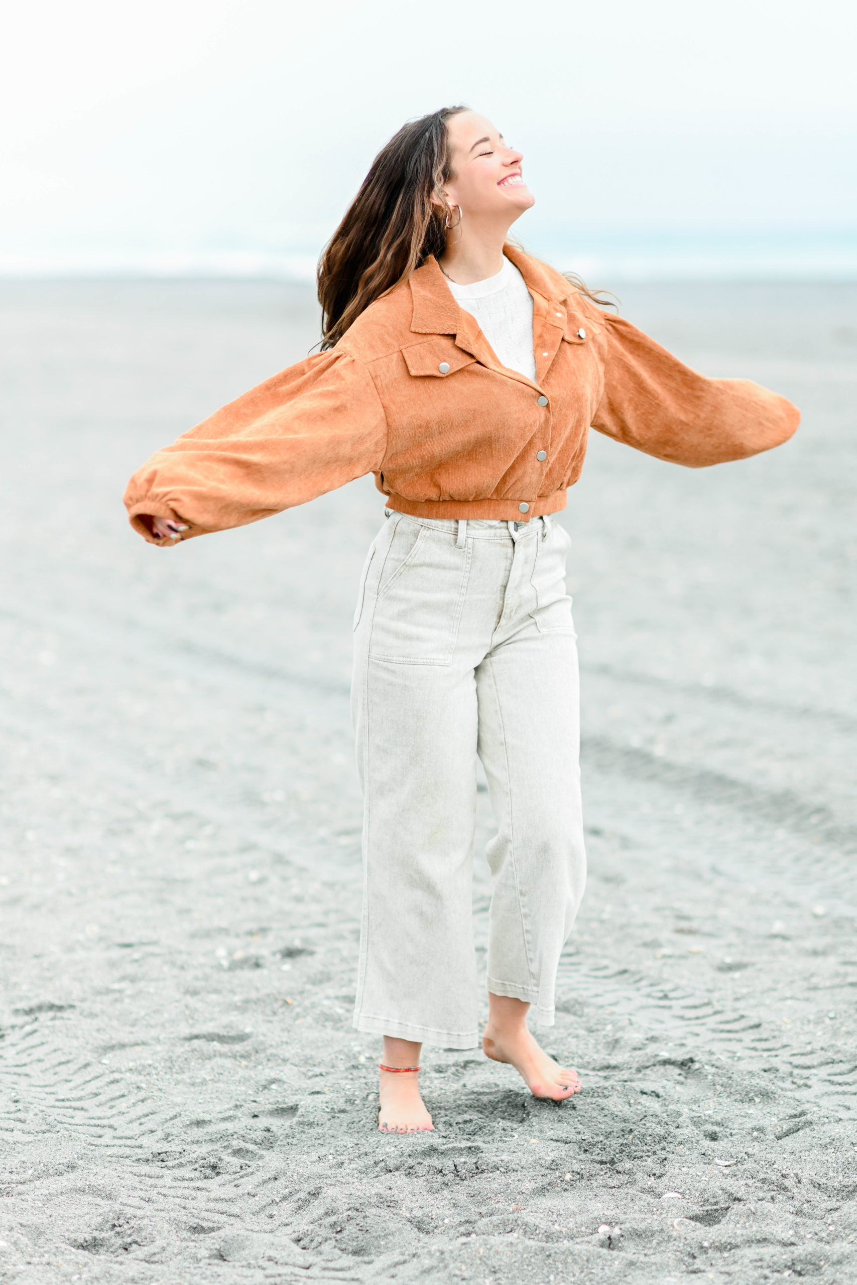 young girl with brown hair in orange jacket and white pants spinning on the beach smiling at the sky