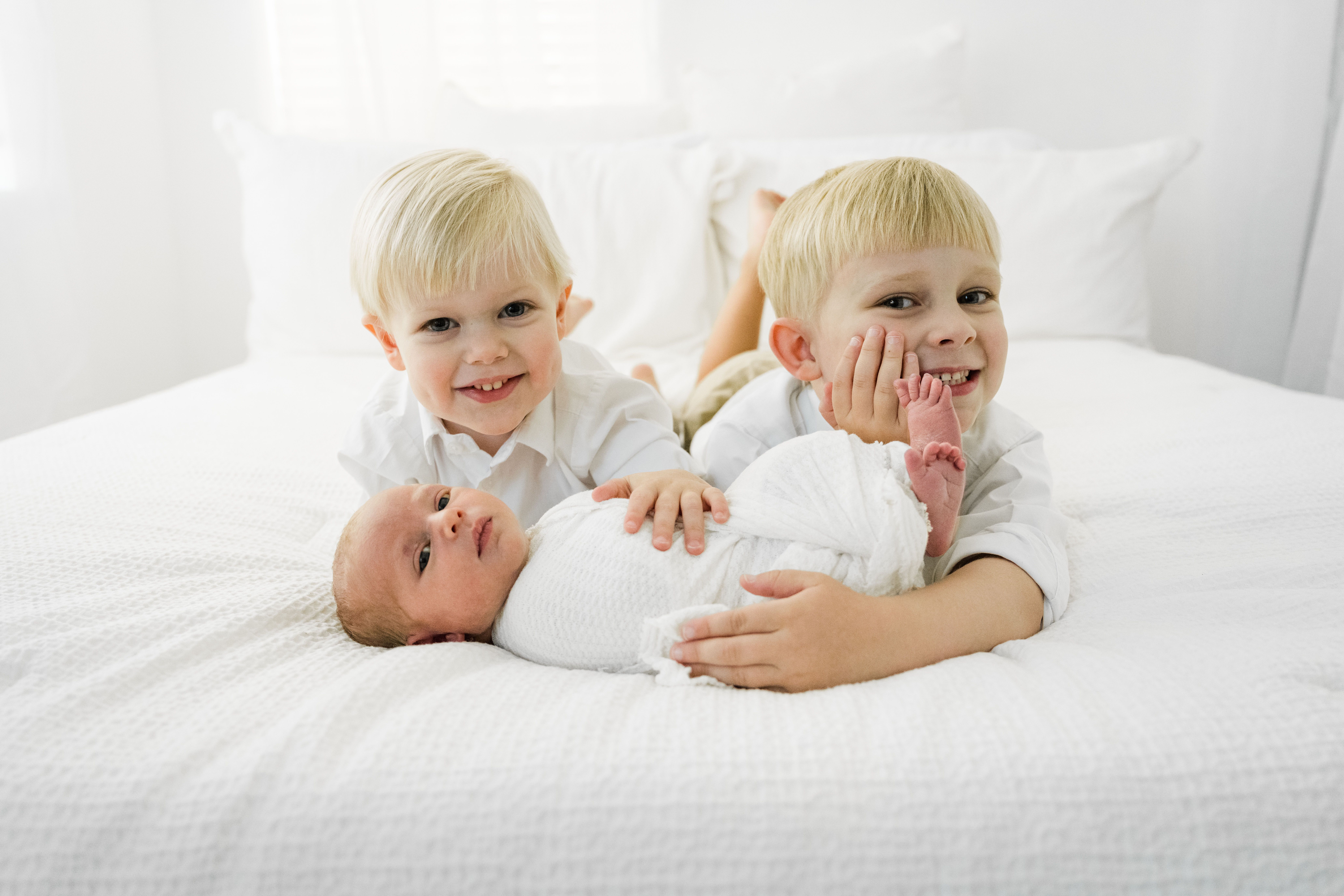 two young boys with their baby sister smile at the camera in this natural light indoors photo session