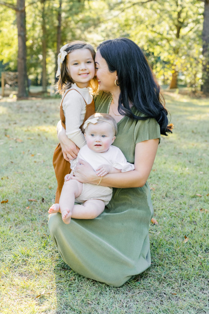 young mom wearing a green dress kneeling in the grass while nuzzling her daughter in a brown jumpsuit and holding her baby in a cream jumper