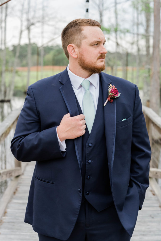 a groom on his wedding day wearing a navy tuxe and green tie staring off into the distance with his right hand holding his lapel and left hand in his pocket
