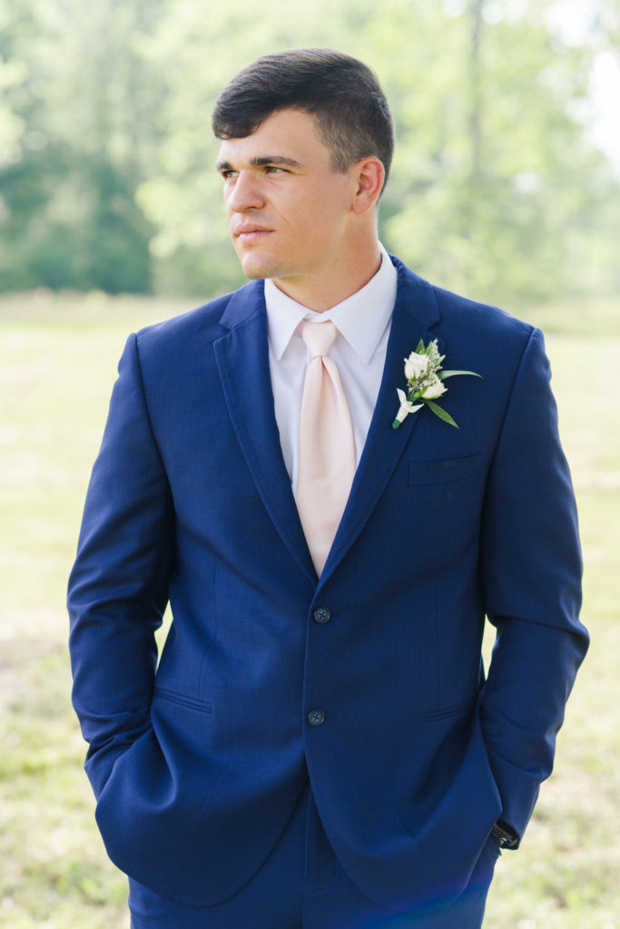a groom on his wedding day wearing a navy tuxe and pink tie staring off into the distance with his hands on his pocket