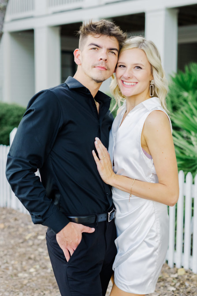 A male wearing an all black outfit poses with his fiance wearing a white short dress pose smiling at the camera while her hand is on his chest and his hands are in his pocket.