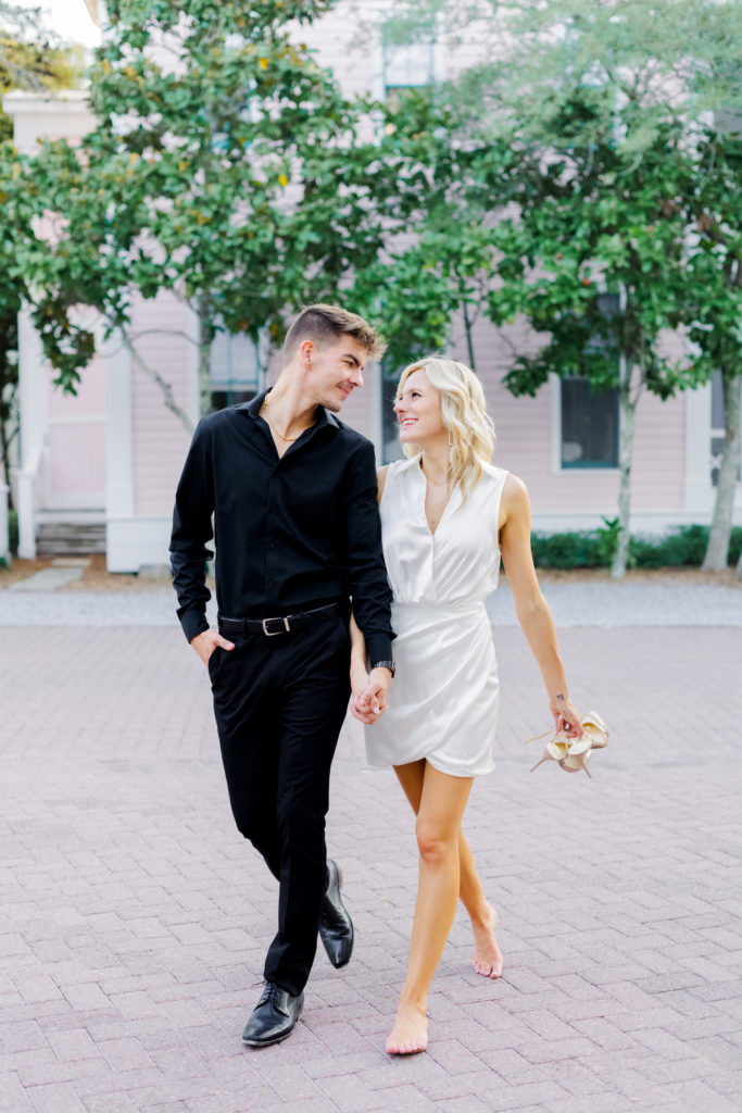 A couple holds hands and walks towards the camera smiling at one another. He is wearing all black and she is wearing a white dress while holding her nude heels. There are green trees and a pink house in the background.