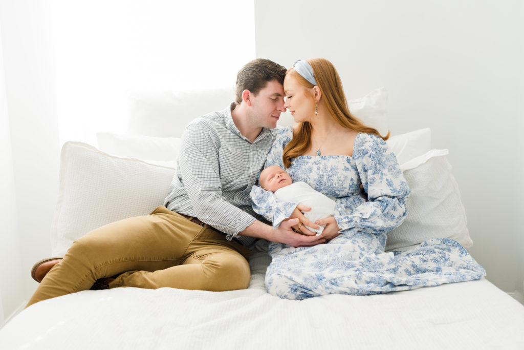 a mother in a blue and white long sleeve dress sits on a white bed with her baby wrapped in a white swaddle as she and her husband who is wearing a blue and green checkered button down and khaki pants look down at their baby