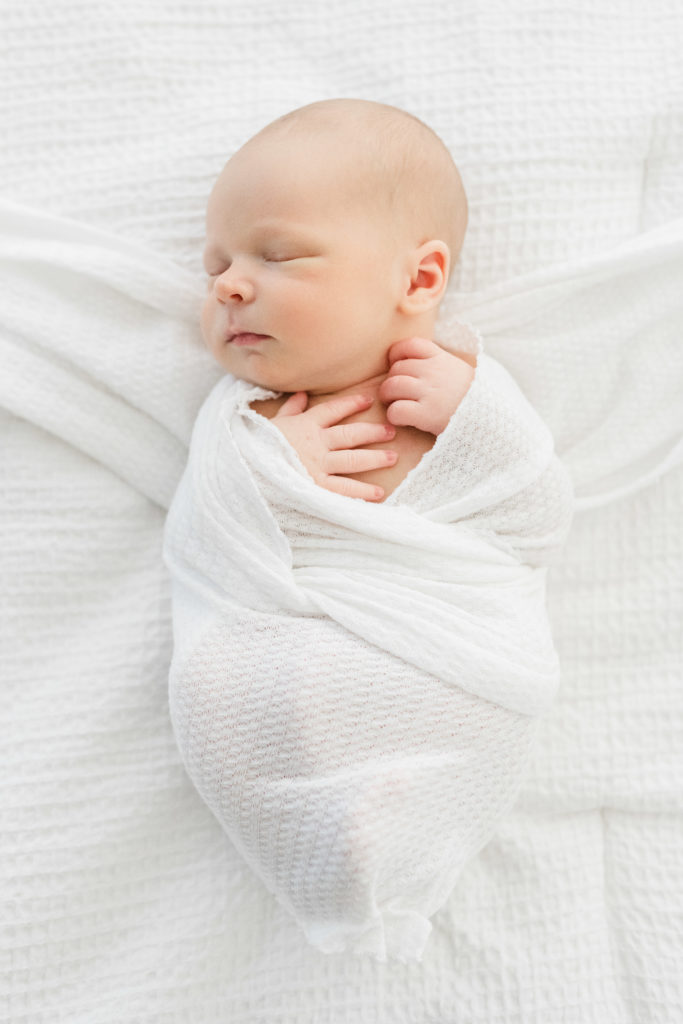 a newborn baby boy swaddled in a white swaddle lays on a white bed with his eyes clothes and hands on his chest