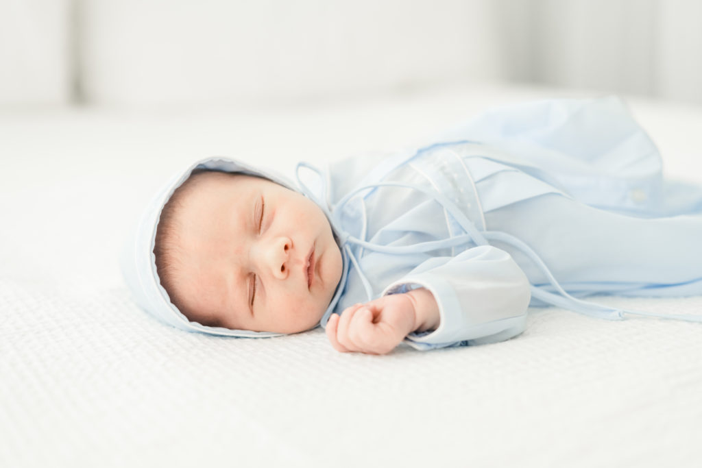 newborn baby boy lays sleeping on white comforter in white studio space wearing a blue gown and bonnet during a newborn session for motherhood photography